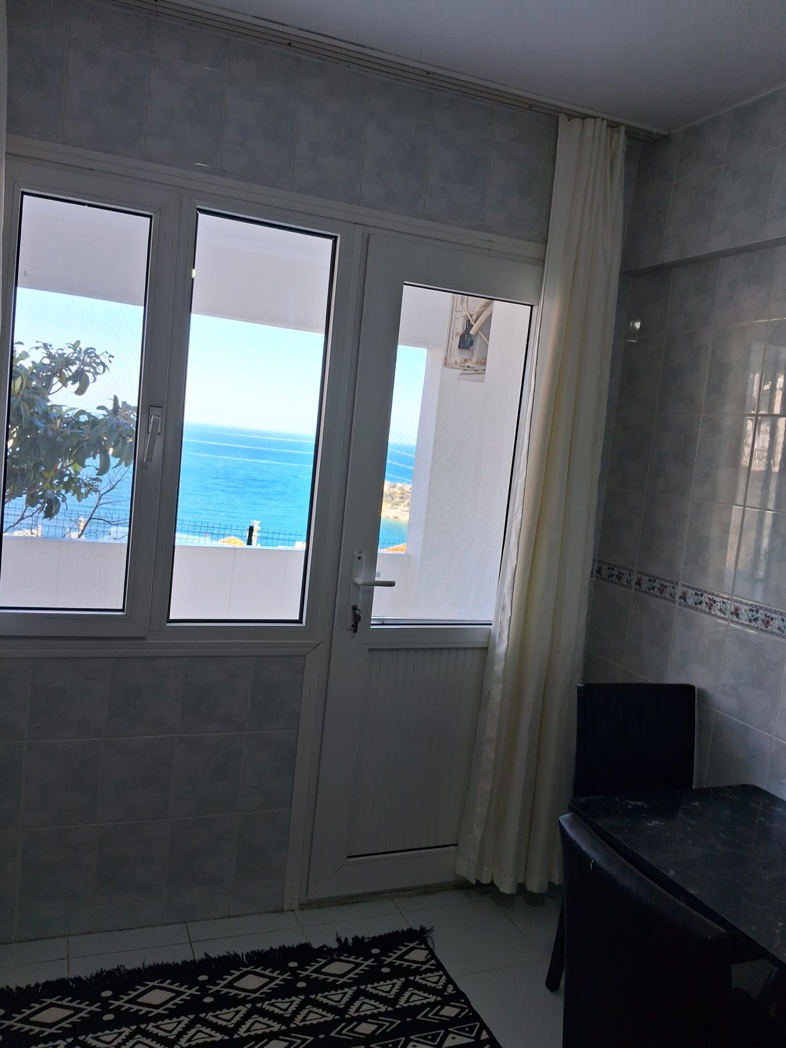 A room with a view of the sea through a glass door, a black cabinet, and a patterned rug.