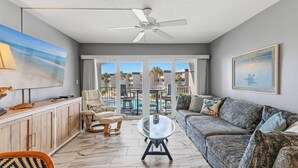 A living room with a sofa, a glass coffee table, a ceiling fan, and a view of a pool and palm trees. - 🦈Great Poolside View | Shuffleboard + Kiddie Pools | Southeaster E203 🌅 (New Smyrna  Beach)