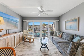 Ein Wohnzimmer mit einer Couch, einem Glastisch, einem Deckenventilator und Blick auf einen Pool und Palmen.