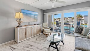 A spacious living area with a ceiling fan, a flat-screen TV, a glass coffee table, a cushioned armchair, and a view of a pool and buildings. - 🦈Great Poolside View | Shuffleboard + Kiddie Pools | Southeaster E203 🌅 (New Smyrna  Beach)