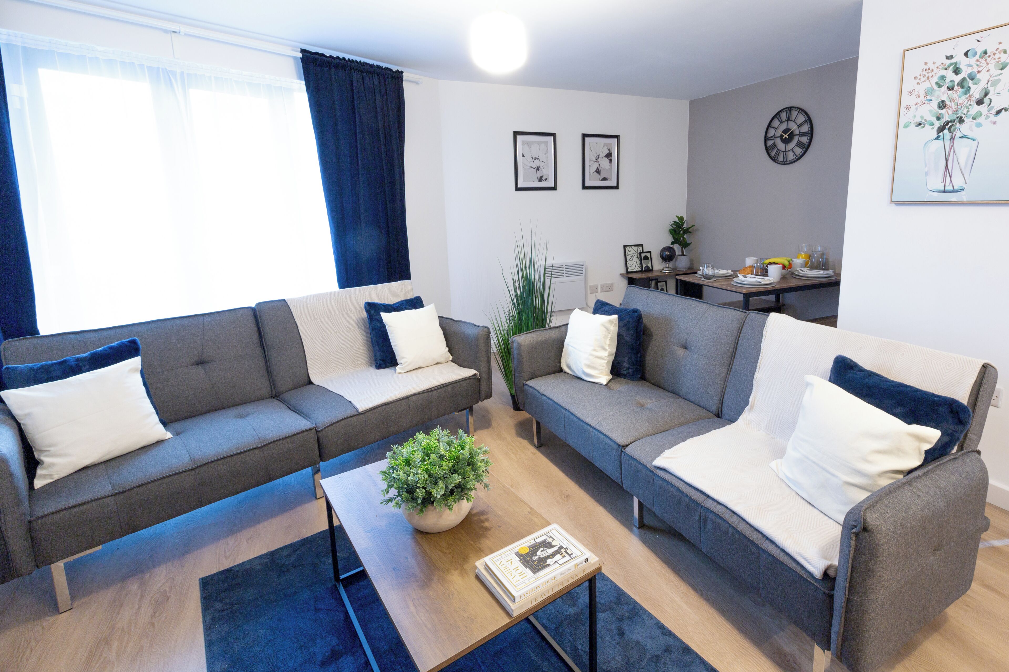 Ein modernes Wohnzimmer mit zwei grauen Sofas, einem Couchtisch und einer kleinen Pflanze. Es gibt einen Essbereich mit einem für das Frühstück gedeckten Tisch.