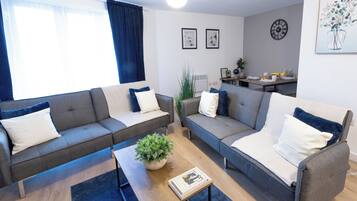 A modern living room with two grey sofas, a coffee table, and a small plant. There is a dining area with a table set for breakfast.