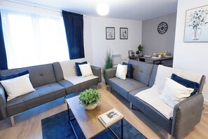 Uma sala de estar moderna com dois sofás cinzentos, uma mesa de centro e uma pequena planta. Há uma área de jantar com uma mesa posta para o café da manhã.