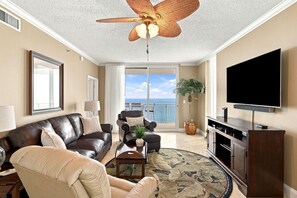 Ein Wohnzimmer mit Deckenventilator, einem großen Flachbildfernseher, Ledersofas, einem Couchtisch und Meerblick durch eine Schiebetür.