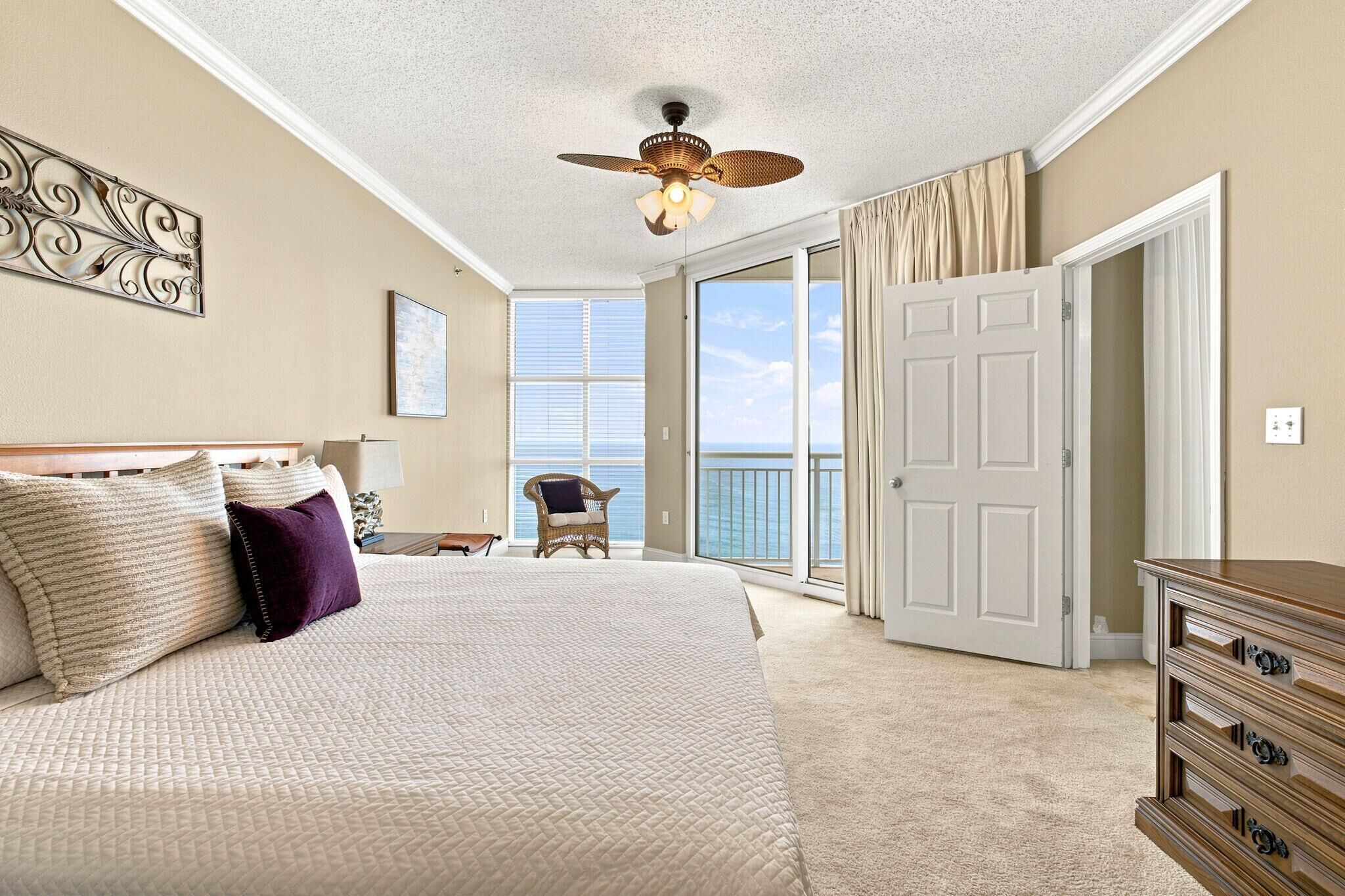 A bedroom with a bed, a dresser, a chair, a ceiling fan, and a view of the ocean through large windows.