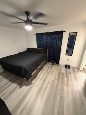 Ein Schlafzimmer mit einem Bett, einem Deckenventilator, einem Fenster mit Vorhängen, einem Spiegel und einem Paar Kurzhanteln auf dem Boden.