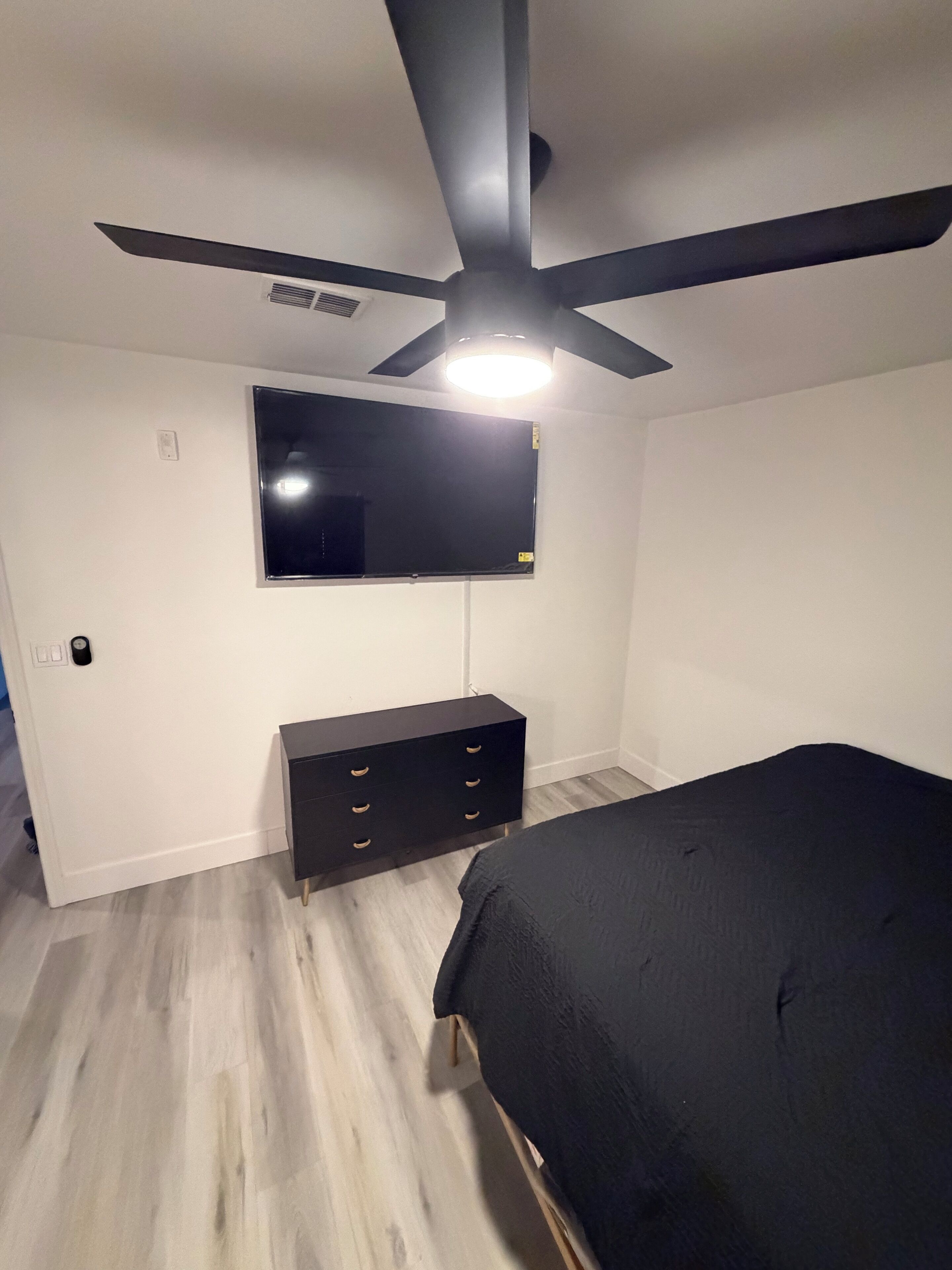 A bedroom with a bed, a dresser, a ceiling fan, and a TV mounted on the wall.