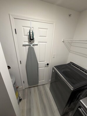 A laundry room with a washer and dryer, a door with a hanging iron, and a white wall. - Delightful 2-bedroom apartment with AC in Summerville (Summerville)
