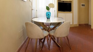 Un comedor con una mesa redonda, cuatro sillas, un florero con una flor, un espejo grande y un televisor sobre un mueble.