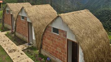 A row of thatched-roof cottages with brick walls, set in a lush valley with mountains in the background.