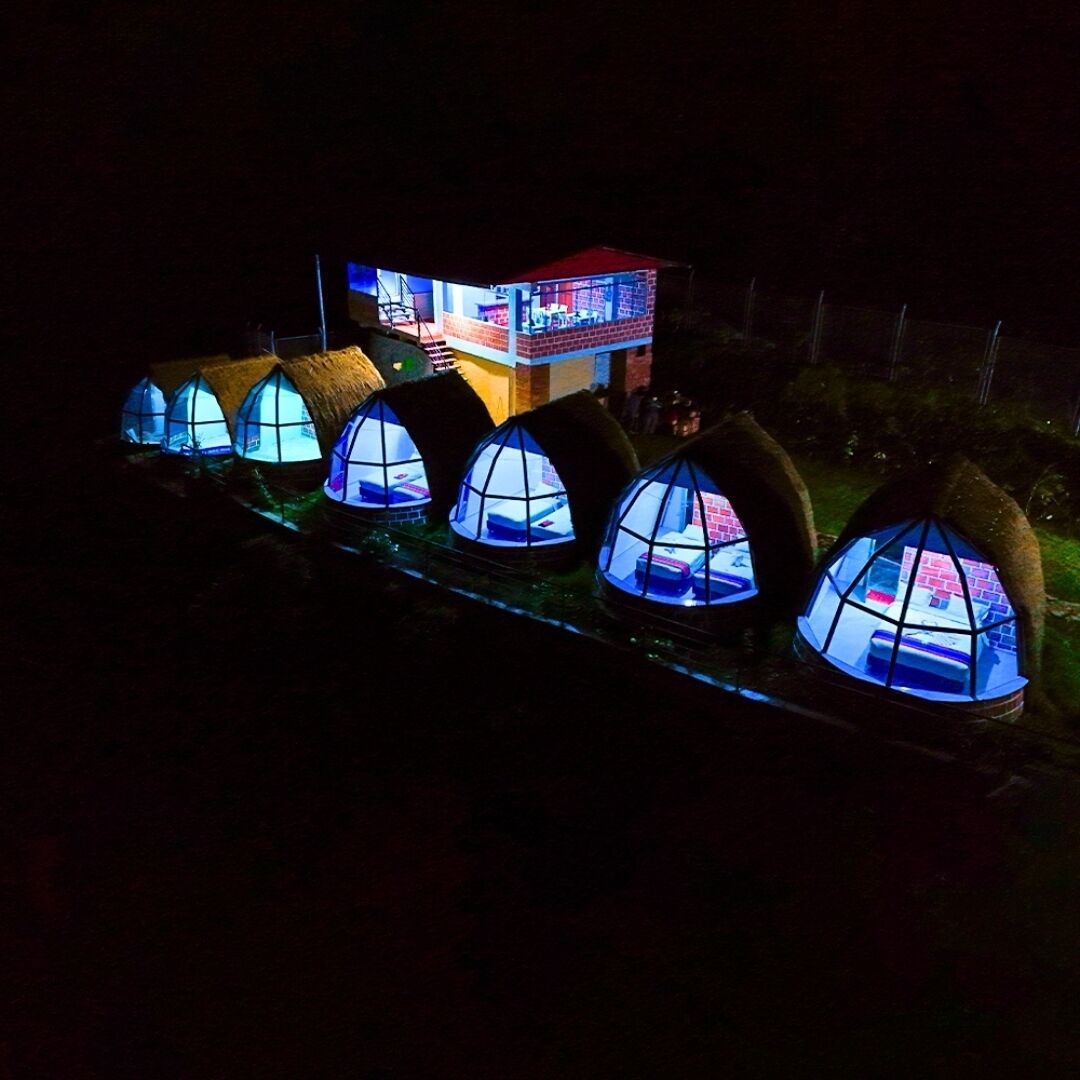 A row of dome-shaped tents with interior lighting, connected by a pathway, and a building with multiple levels in the background.