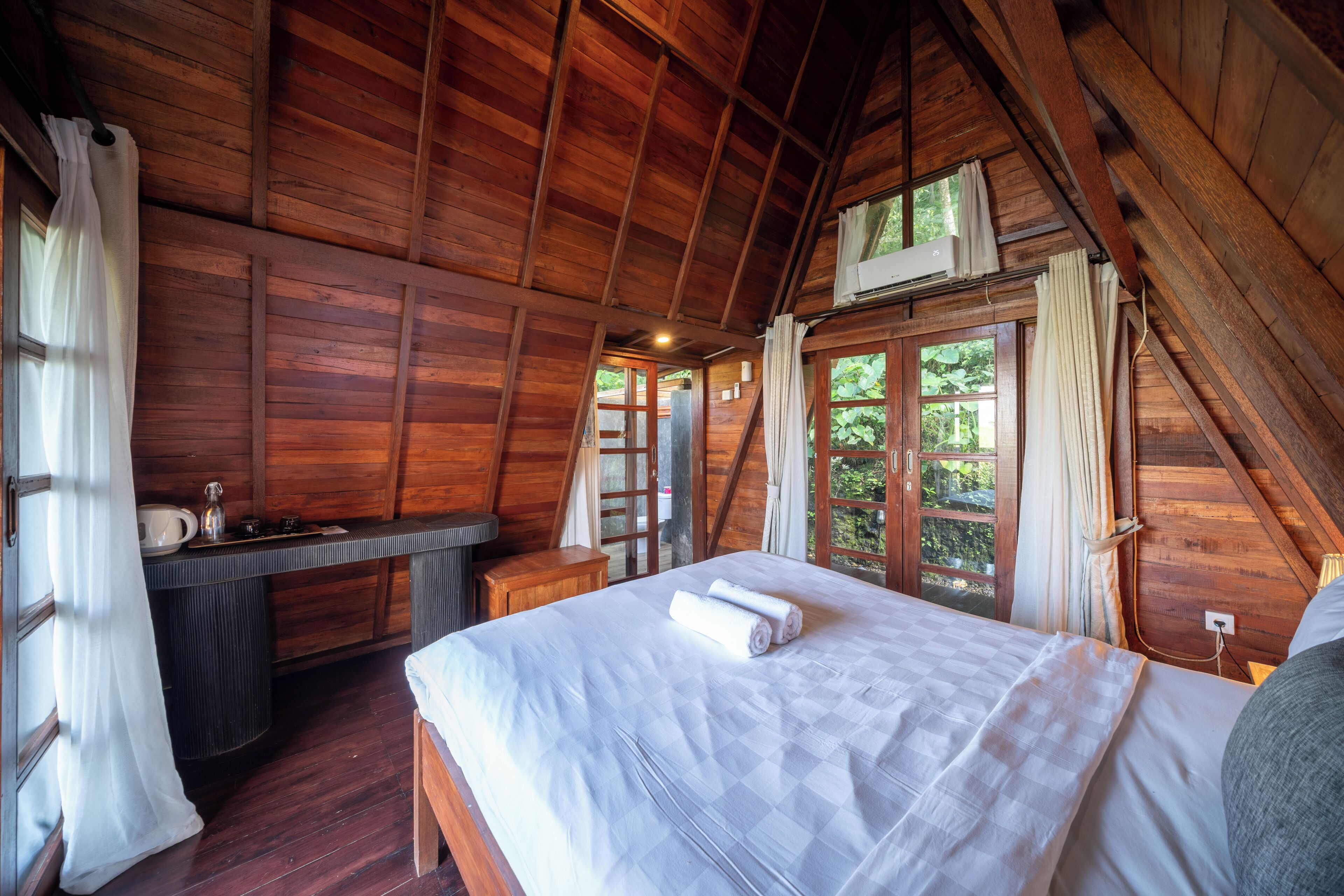 Een slaapkamer in een houten hutje, met een bed, een kleine tafel en een raam met uitzicht op groen.