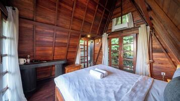 Een slaapkamer in een houten hutje, met een bed, een kleine tafel en een raam met uitzicht op groen.