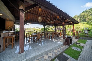 Une terrasse extérieure avec des tables et des chaises en bois, un espace bar et une vue sur une végétation luxuriante.