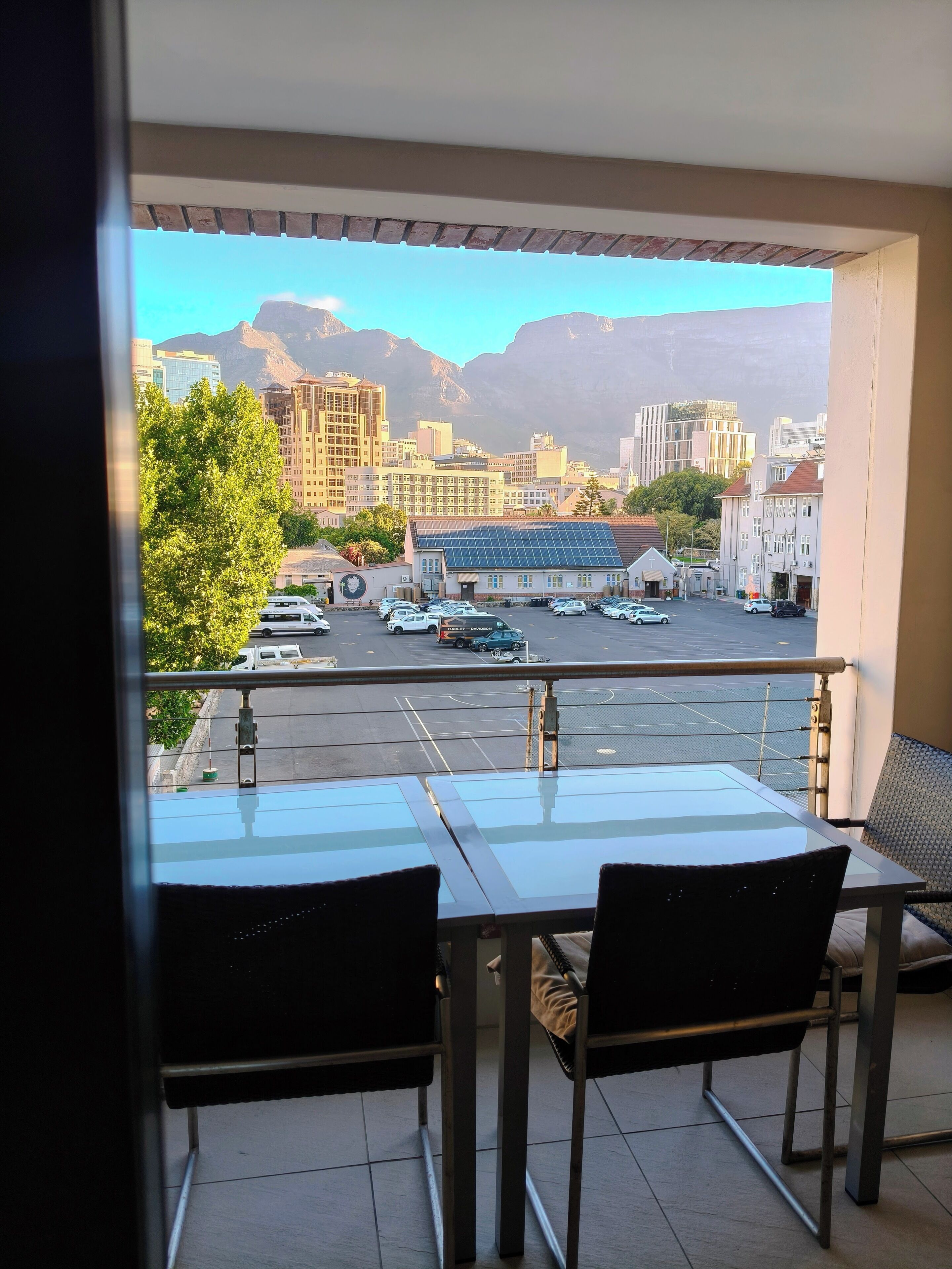 Un balcón con mesa y sillas, con vistas a un estacionamiento y edificios, con montañas al fondo.