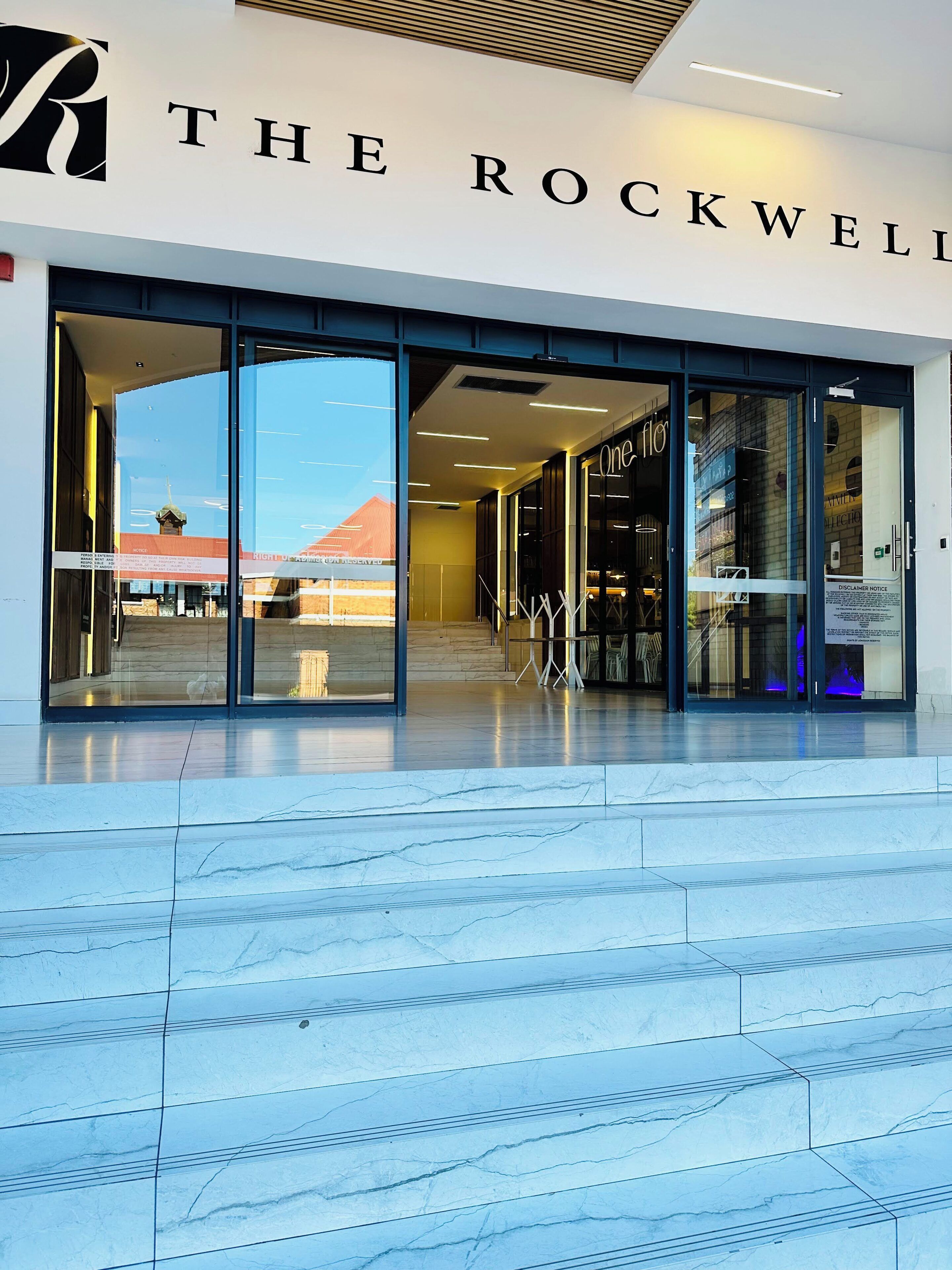 A modern building entrance with glass doors, a sign reading 'THE ROCKWELL,' and a clear view of the sky and a distant building.