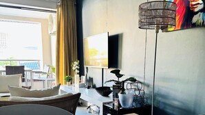 A modern living room with a wooden dining table, a sofa, a TV mounted on the wall, and a large painting. - Stylish 2-Bed Apartment with Pool, Gym & Mountain Views. Walk to V&A Waterfront! (Cape Town)