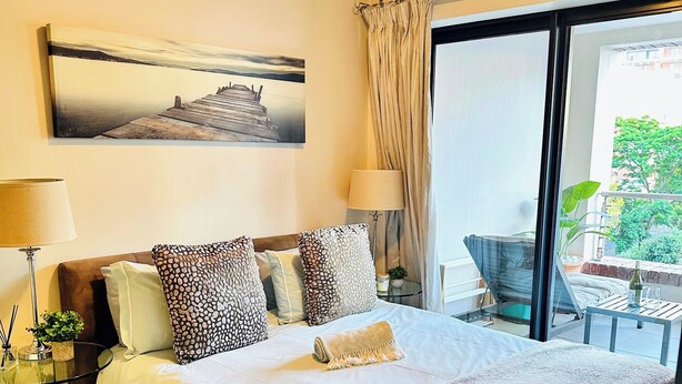 A hotel room with a large bed, a painting on the wall, a lamp, and a view of the outdoors. - Stylish 2-Bed Apartment with Pool, Gym & Mountain Views. Walk to V&A Waterfront! (Cape Town)
