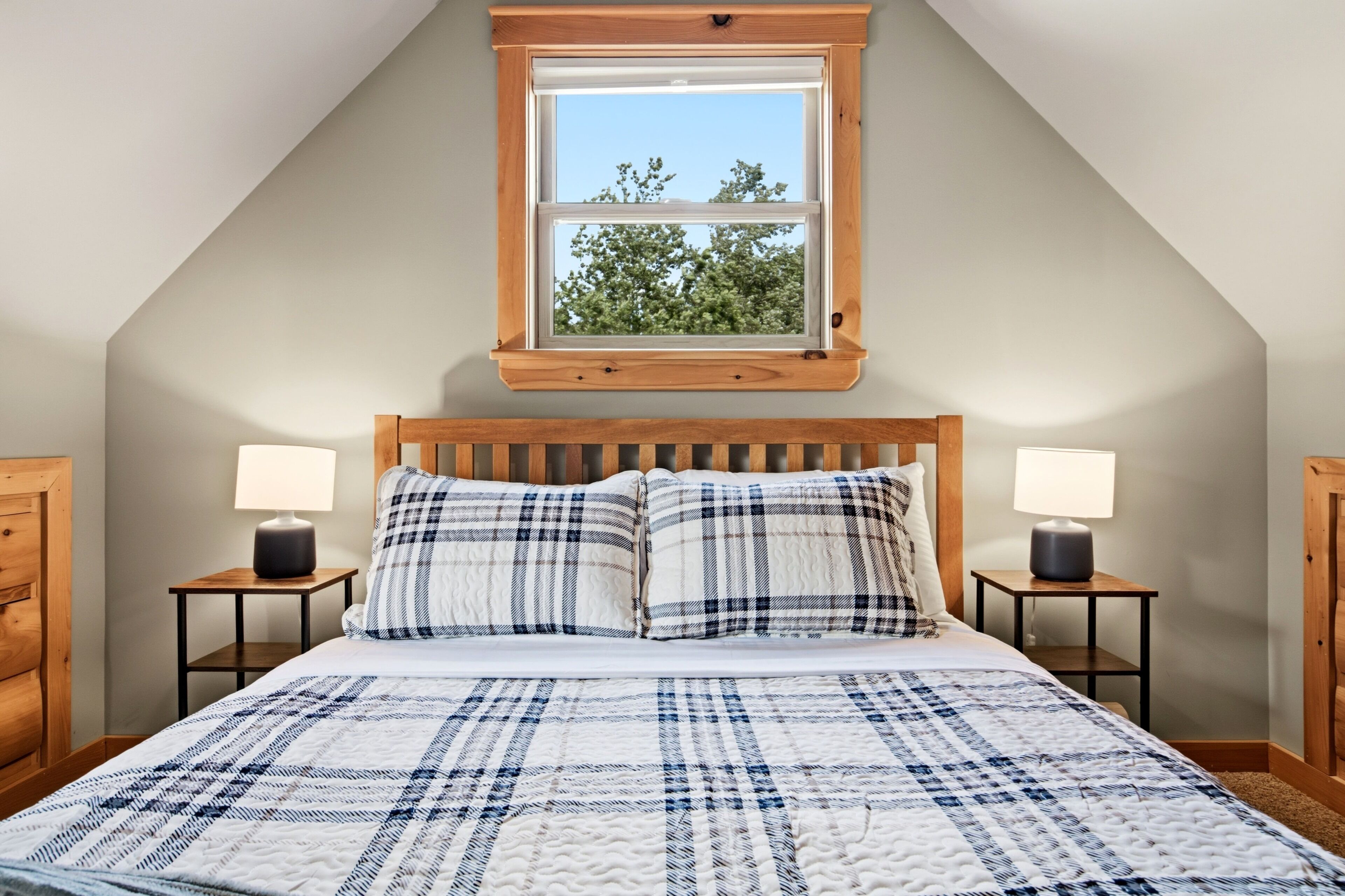 Un dormitorio con techo inclinado, cabecera de madera, cama con colcha azul y blanca a cuadros, dos lámparas de noche y una ventana con vistas a árboles.