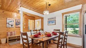 A wooden-paneled dining room with a large table set for six, a bench with plaid cushions, and a large window with a view of greenery. - Peak and Pine Hideway / Hot Tub & Arcade (Bethel)