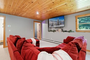 A living room with a wooden ceiling, a mounted flat-screen TV, red sofas, and a window with a view of trees. - Peak and Pine Hideway / Hot Tub & Arcade (Bethel)