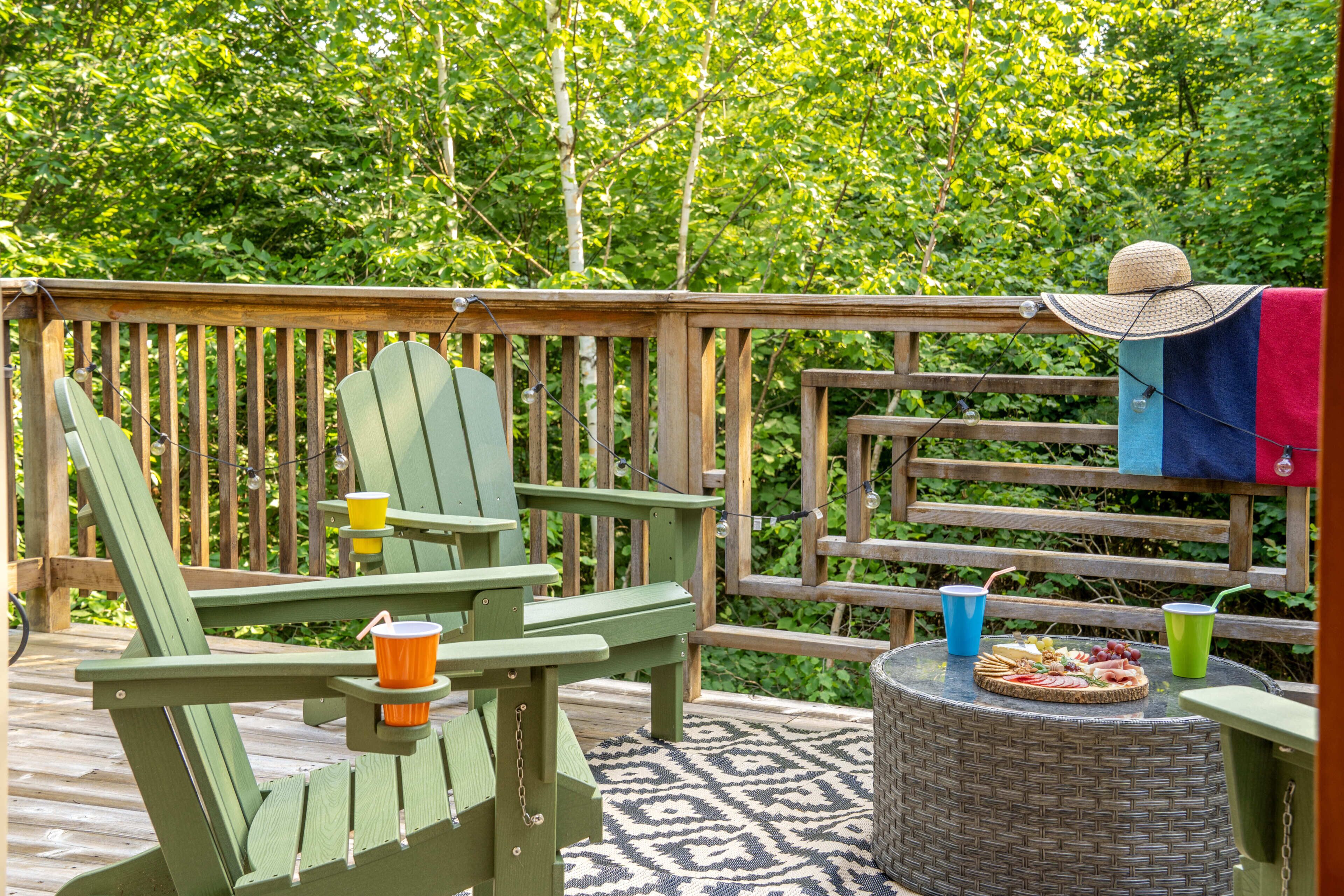A wooden deck with Adirondack chairs, a wicker table with food and drinks, and a hat on a chair.