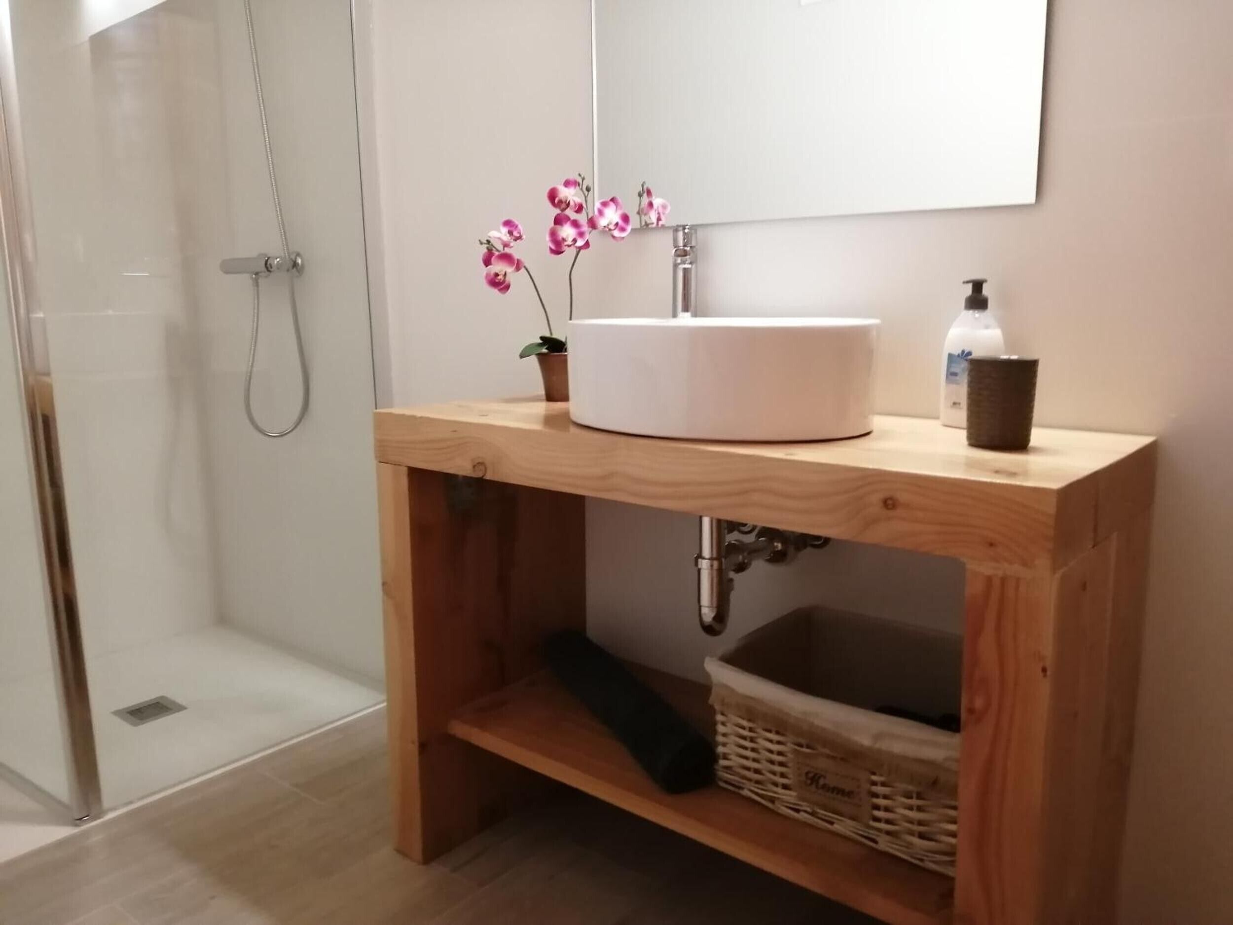 A bathroom with a wooden vanity, a white sink, a mirror, a shower, and a small potted orchid.
