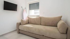 En beige soffa i ett rum med en platt-TV som är monterad på väggen, ett fönster med persienner och ett klinkergolv.