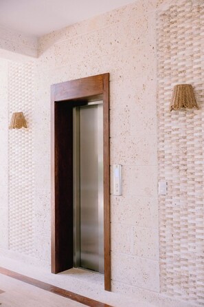 A modern elevator with a wooden frame and a metallic interior, set in a hallway with textured walls and a woven light fixture. - Casa Playa, Las Iguanas CapCana (Punta Cana)