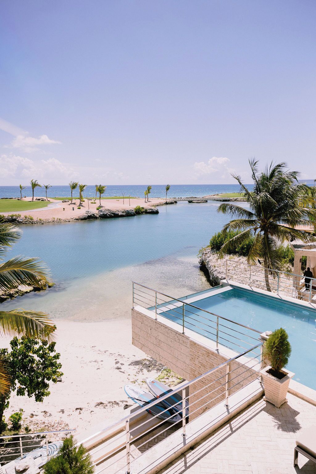 Une piscine avec vue sur la plage et l’océan, entourée de palmiers et surplombée par un petit pont.