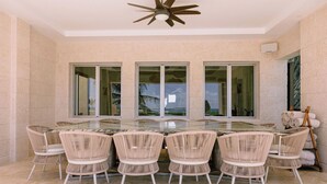 A dining area with a glass-top table surrounded by wicker chairs, large windows, and a ceiling fan. - Casa Playa, Las Iguanas CapCana (Punta Cana)