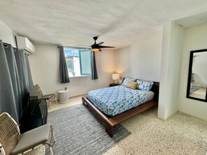 Un dormitorio con cama, ventilador de techo, ventana con cortinas, mesita de noche con lámpara y una silla.