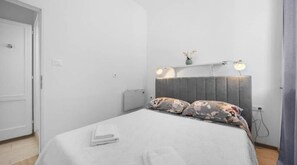 Ein modernes Schlafzimmer mit grauem Kopfteil, zwei Nachttischlampen, einem blumigen Kopfkissen und einem ordentlich bezogenen Bett.
