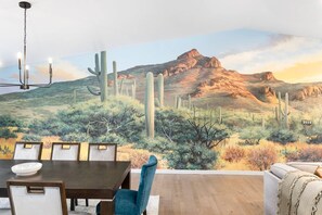 A dining area with a table and chairs, a sofa, and a mural of a desert landscape with cacti and a mountain. - Epic PHX Fun-Game Room•Pool•Hot Tub•Mtn Views (Phoenix)