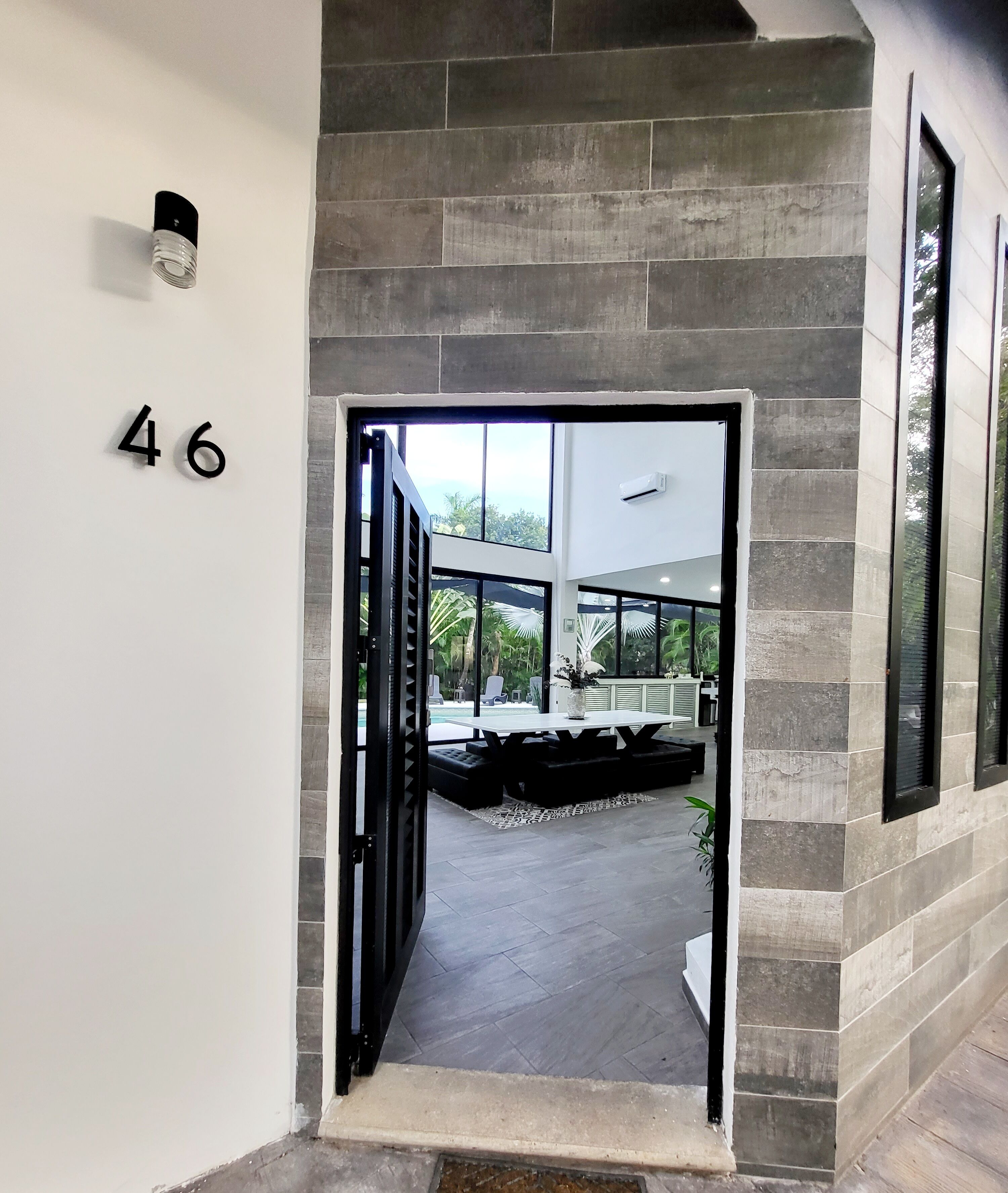 Un couloir moderne, avec le numéro « 46 » affiché sur le mur, mène à un patio doté d’une piscine, de mobilier d’extérieur et offrant une vue sur la verdure.
