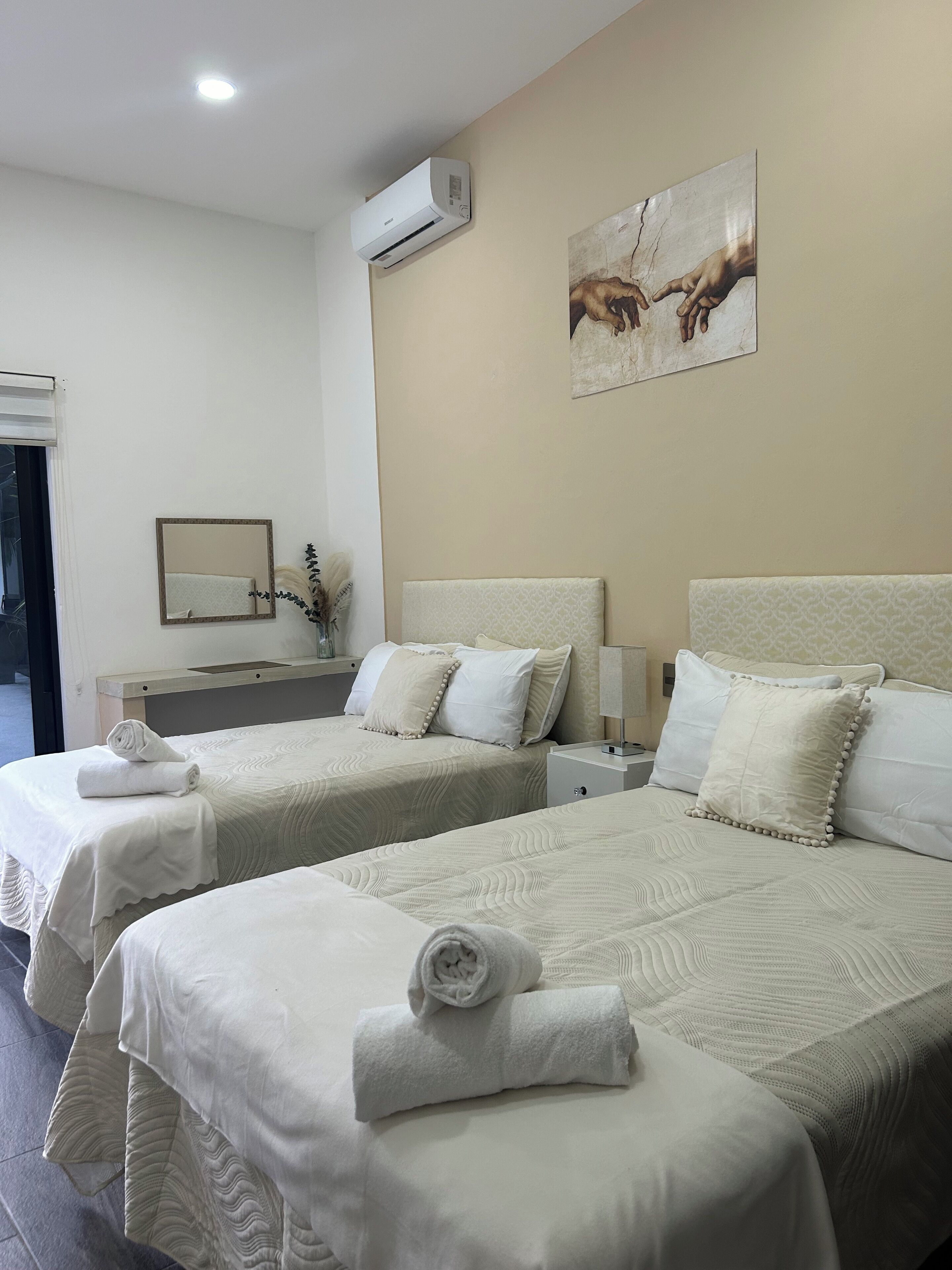 A hotel room with two beds, a wall-mounted air conditioner, a painting, a mirror, and a bedside table with a lamp.