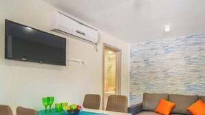 Ein Wohnzimmer mit Flachbildfernseher, einem Esstisch mit Stühlen und einer Couch mit orangenen Kissen.