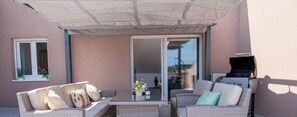 Une terrasse extérieure aménagée avec des meubles en osier, un barbecue et une porte-fenêtre coulissante donnant sur un espace intérieur.