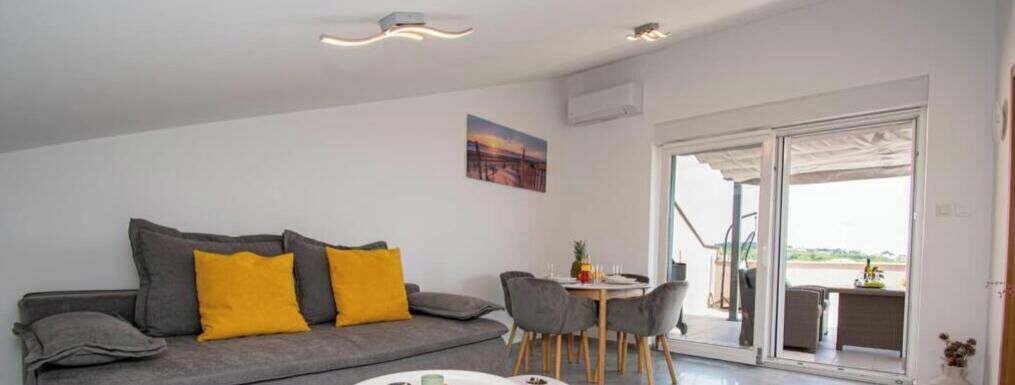 Ein modernes Wohnzimmer mit einem grauen Sofa, gelben Kissen, einem Esstisch mit Stühlen und einer großen Glasschiebetür zu einem Außenbereich.