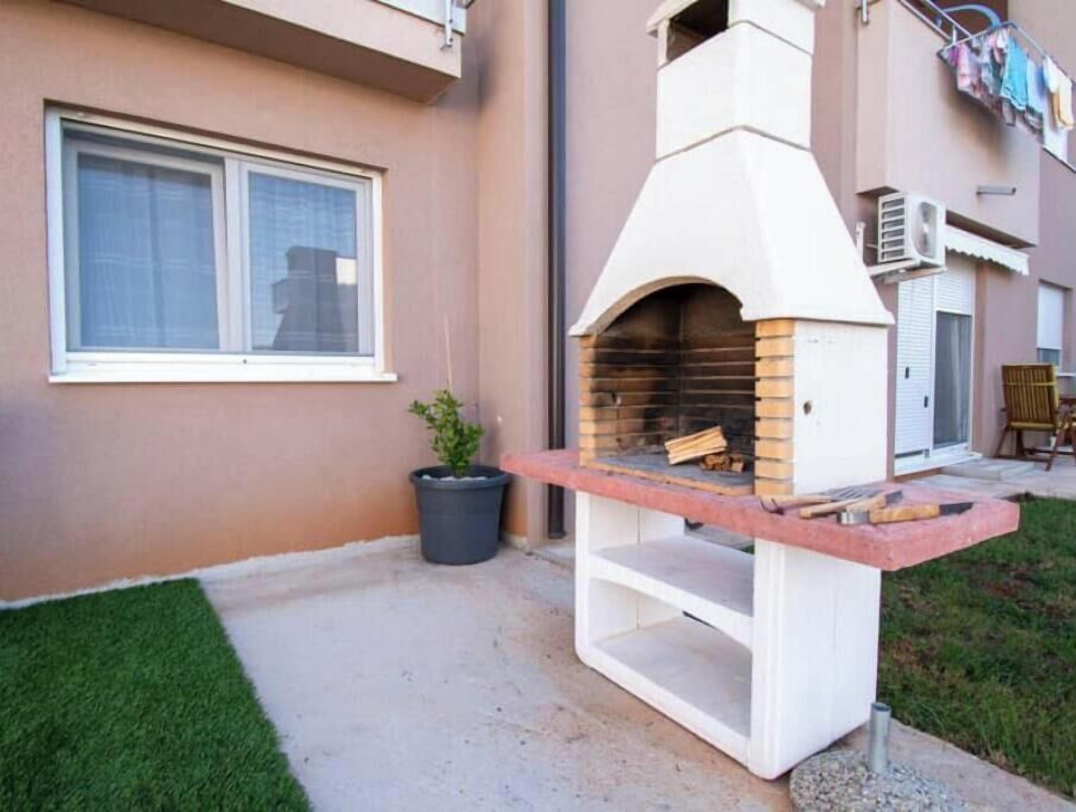 Zona de barbacoa al aire libre con horno de ladrillo, chimenea blanca, planta en maceta y ventana con persianas.
