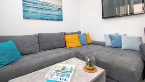 Um sofá modular cinza com almofadas azuis e amarelas, uma mesa de centro com tampo de vidro e uma revista, e uma pintura com uma paisagem oceânica azul na parede.