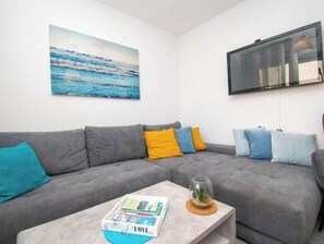 Ein graues Ecksofa mit blauen und gelben Kissen, ein Couchtisch mit Glasplatte und einer Zeitschrift, sowie ein Wandbild mit einer Ansicht des blauen Ozeans.