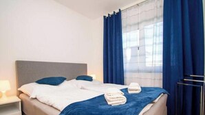 Cama bem arrumada com travesseiros e um cobertor azul, mesa de cabeceira com abajur e janela com cortinas azuis.