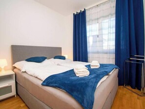 Ein ordentlich gemachteres Bett mit Kissen und einer blauen Decke, ein Nachttisch mit einer Lampe sowie ein Fenster mit blauen Vorhängen.
