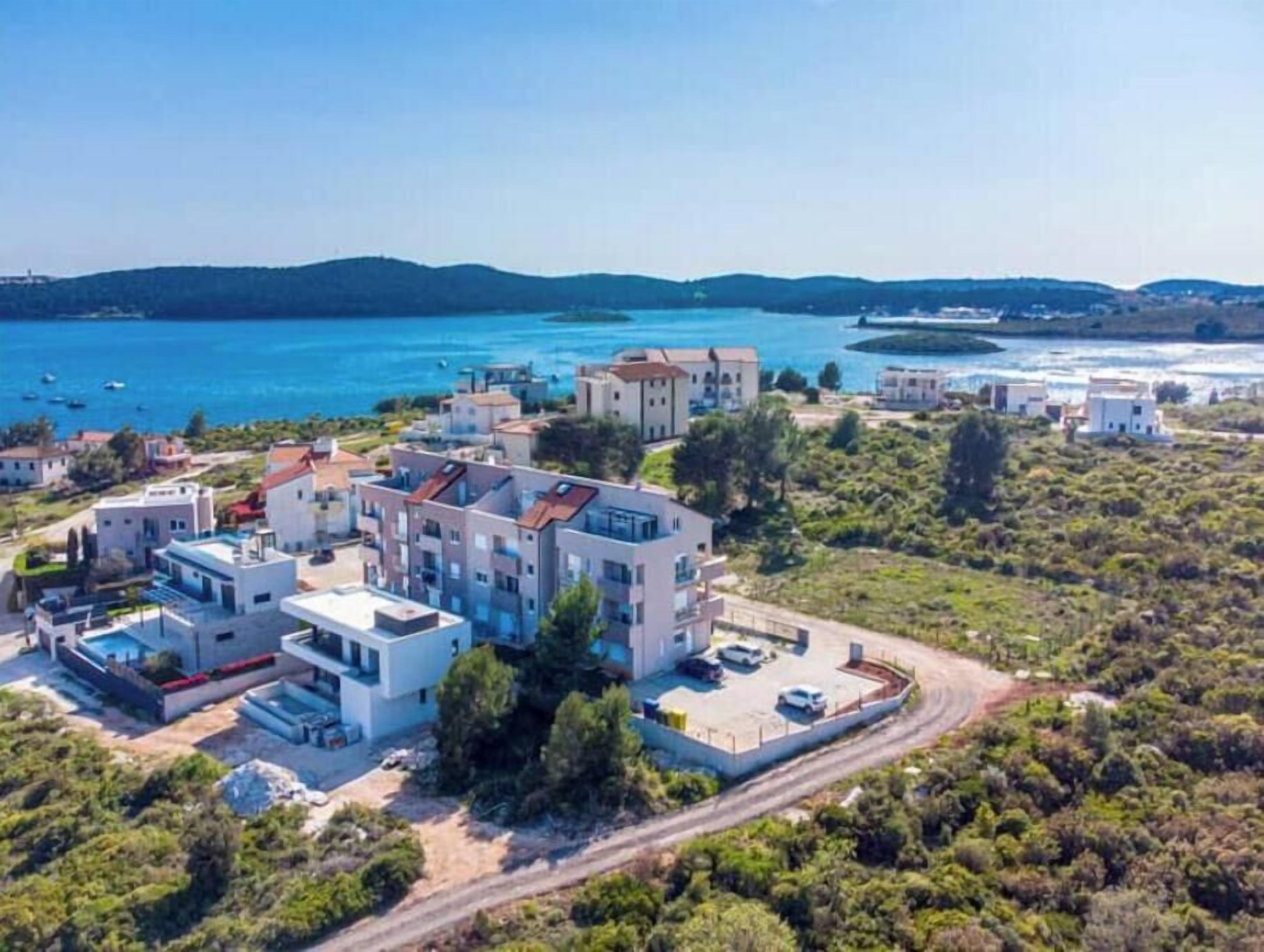 A coastal residential area with modern buildings, a clear blue sea, and a hilly landscape in the background.