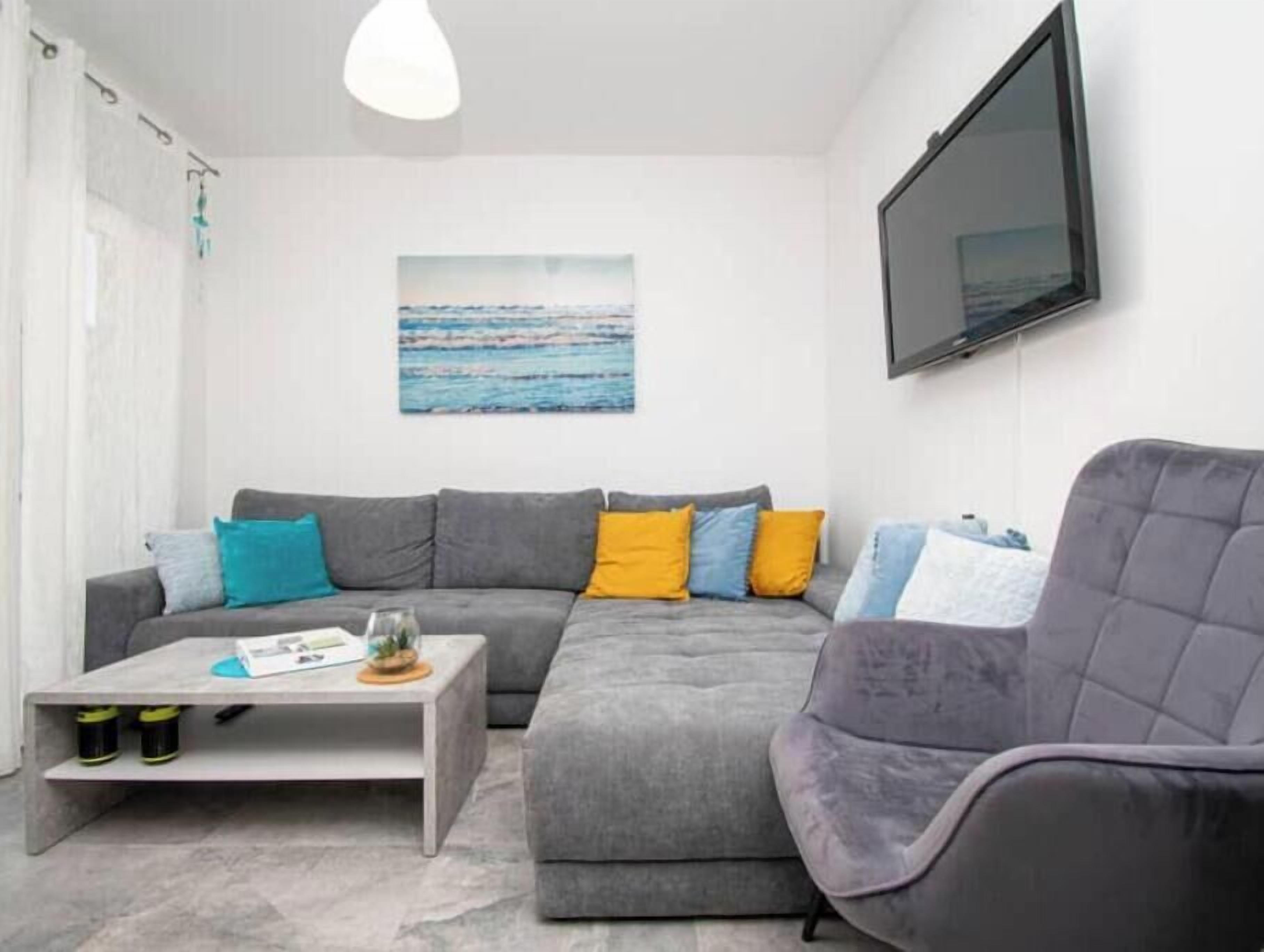 Een moderne woonkamer met een grijze modulaire bank, een witte salontafel en een grijze fauteuil. Een flatscreen televisie is aan de muur boven de bank bevestigd.