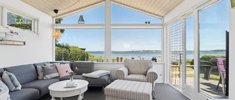 A sunlit room with a grey sofa, a striped ottoman, and a round coffee table. Large windows offer a view of the sea and greenery.