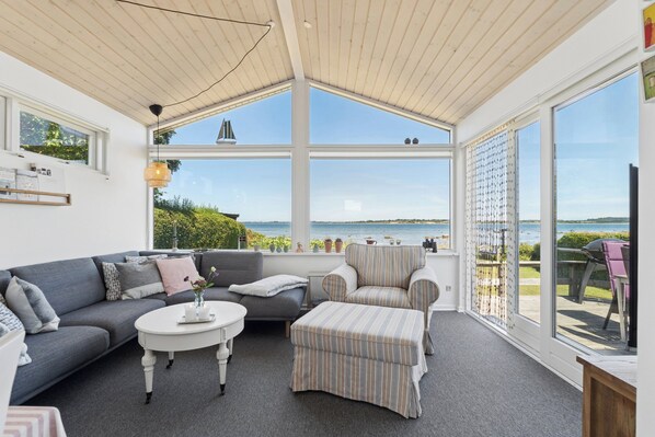 A sunlit room with a grey sofa, a striped ottoman, and a round coffee table. Large windows offer a view of the sea and greenery.