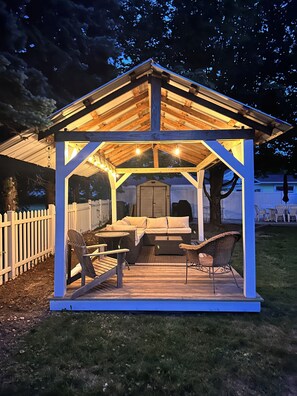 Ein hölzerner Gazebo mit einer Bank, Stühlen und einem Tisch, sowie einem überdachten Bereich mit Lichterketten.