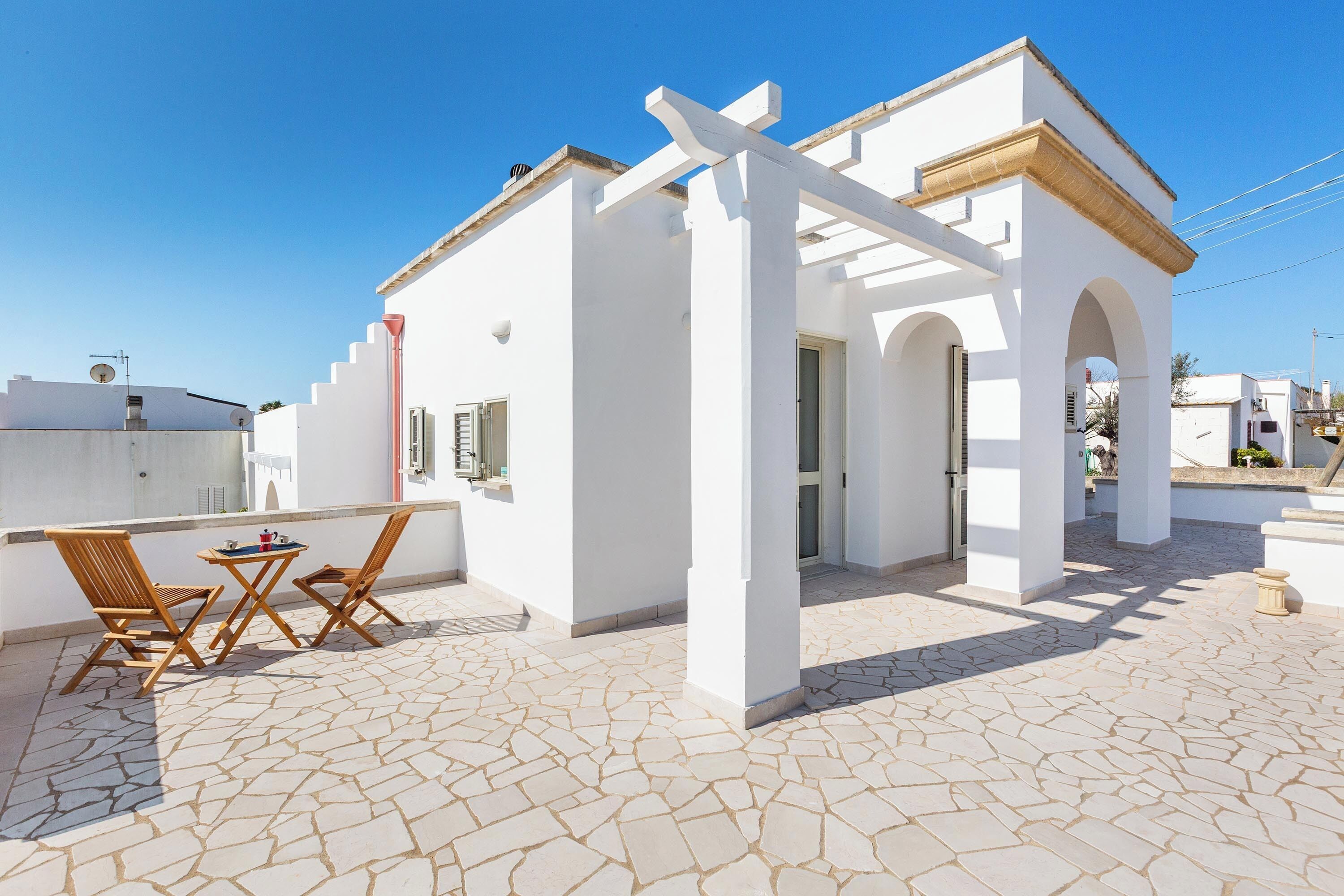 A white building with a tiled patio, wooden chairs, and a table.